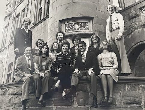 A group of thirteen people pose for a photo on stone steps outside a large building with ornate windows.