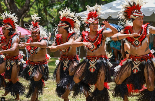 A group of performers in traditional attire dancing outdoors on a grassy area. Their costumes feature ornate headpieces with white and red feathers, geometric-patterned necklaces, and black skirts with red and white accents.