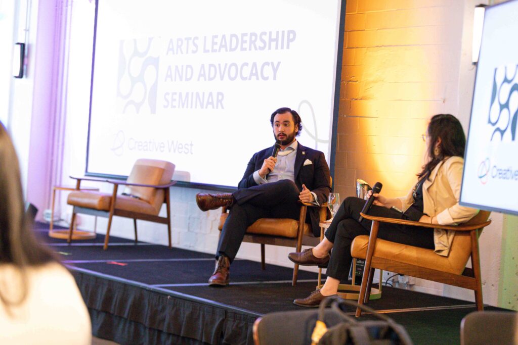 Two people seated on a stage holding microphones. The screen behind speakers reads "Arts Leadership and Advocacy Seminar"