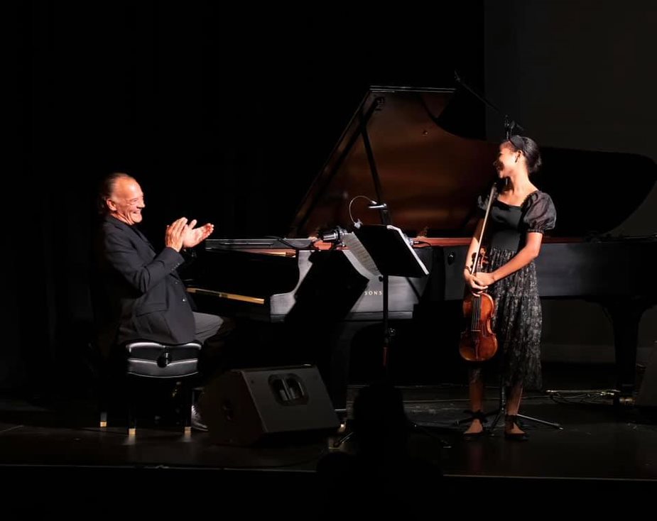 A man seated at a grand piano claps while a woman with a violin stands on stage, both smiling.