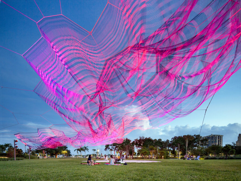A pink net-like installation suspended over a park with people sitting on the grass, set against a clear blue sky and palm trees.