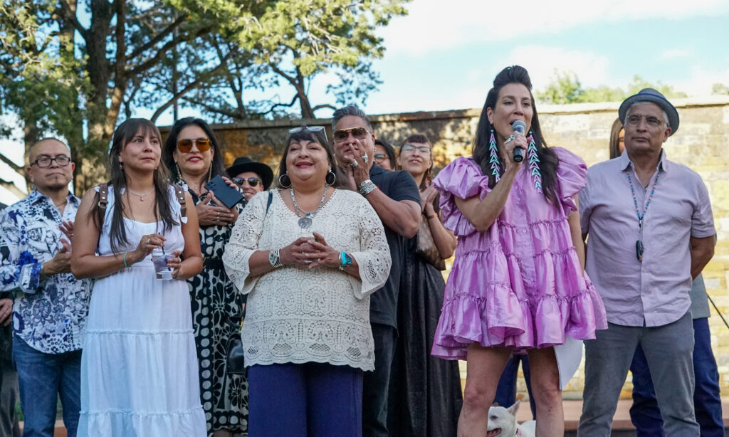 A group of people stand on an outdoor stage; a person in a dress speaks into a microphone while a dog stands at their feet.