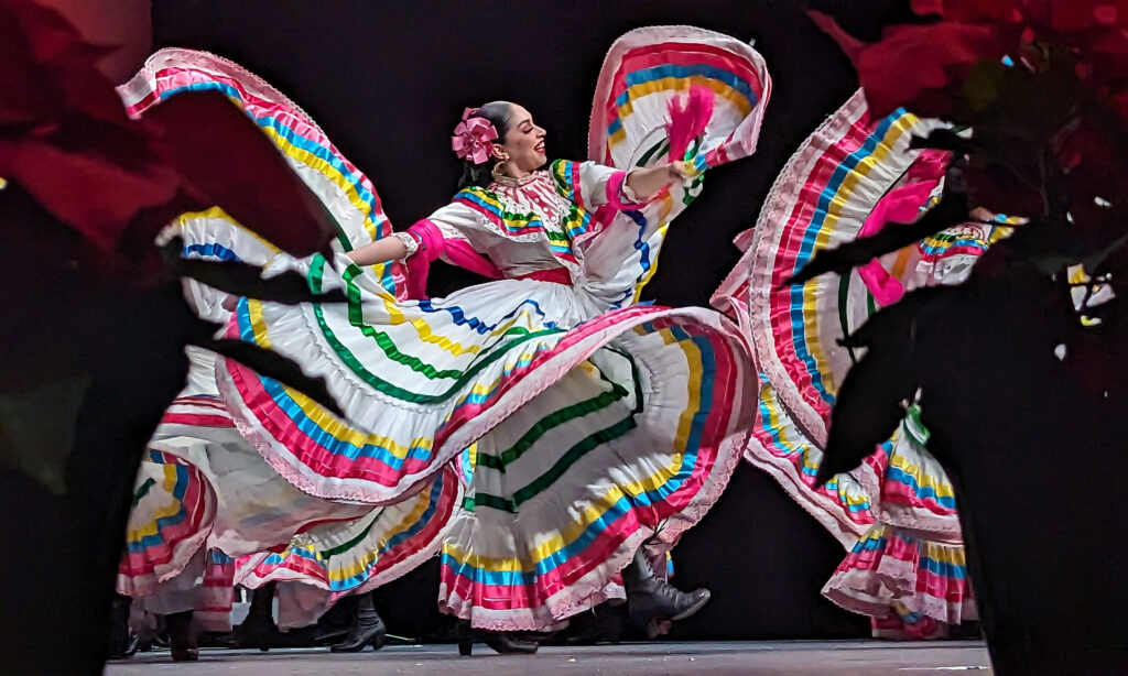 A group dancing in a vibrant, traditional Mexican dress with colorful stripes and ruffled edges, captured mid-dance. They are smiling, with their hair styled in a bun adorned with flowers.