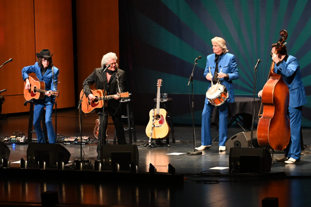 Four musicians performing on stage, wearing blue and black suits, playing guitars, a snare drum, and an upright bass.