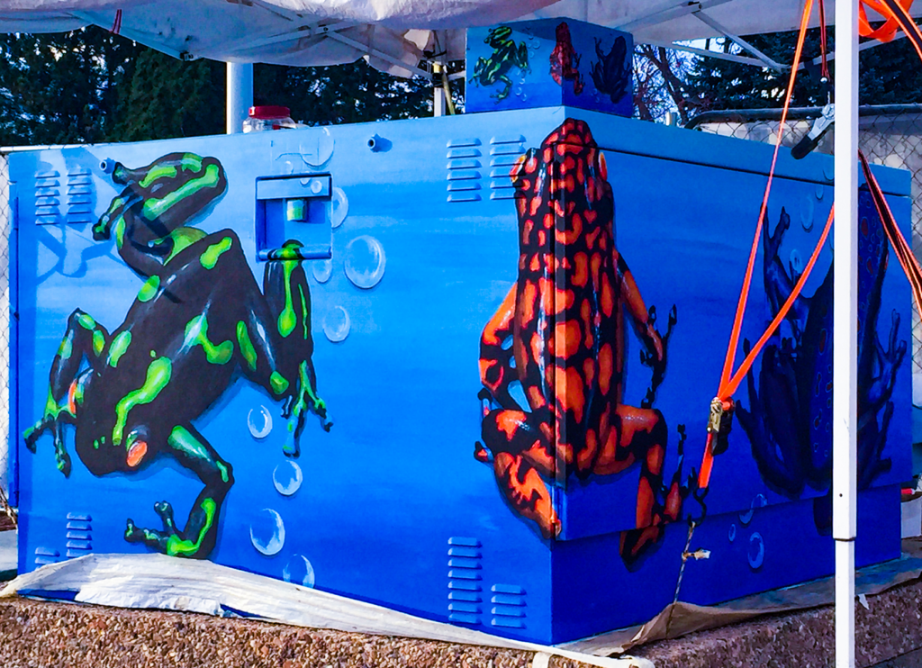 A brightly painted utility box featuring frogs in vivid green, black, red, and orange against a blue background with bubbles.