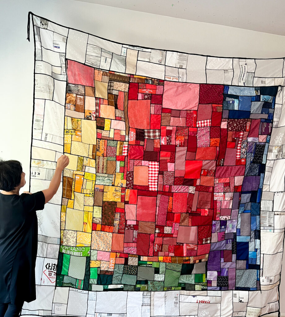 A colorful quilt hung up on a white wall and a woman standing next to it.