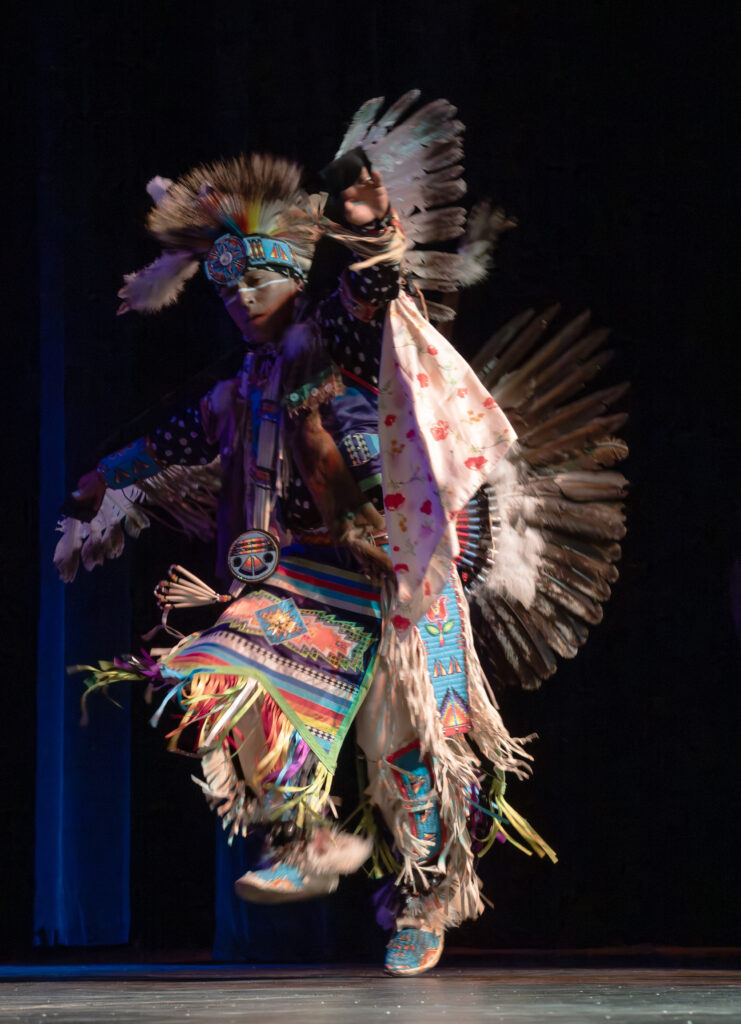 A person performing a traditional dance in vibrant regalia, featuring a feathered headdress, floral-patterned shawl, and intricate beadwork. Their dynamic pose highlights the colorful attire, with a dark background emphasizing the movement and cultural significance of the performance