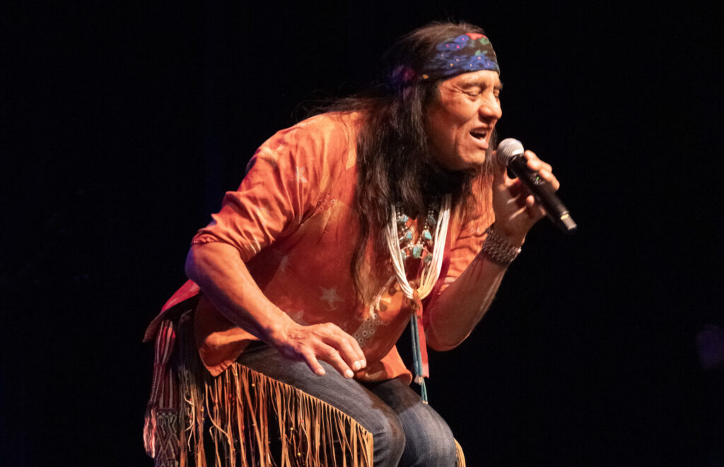 An Indigenous performer standing on a stage under the spotlight holding a microphone.