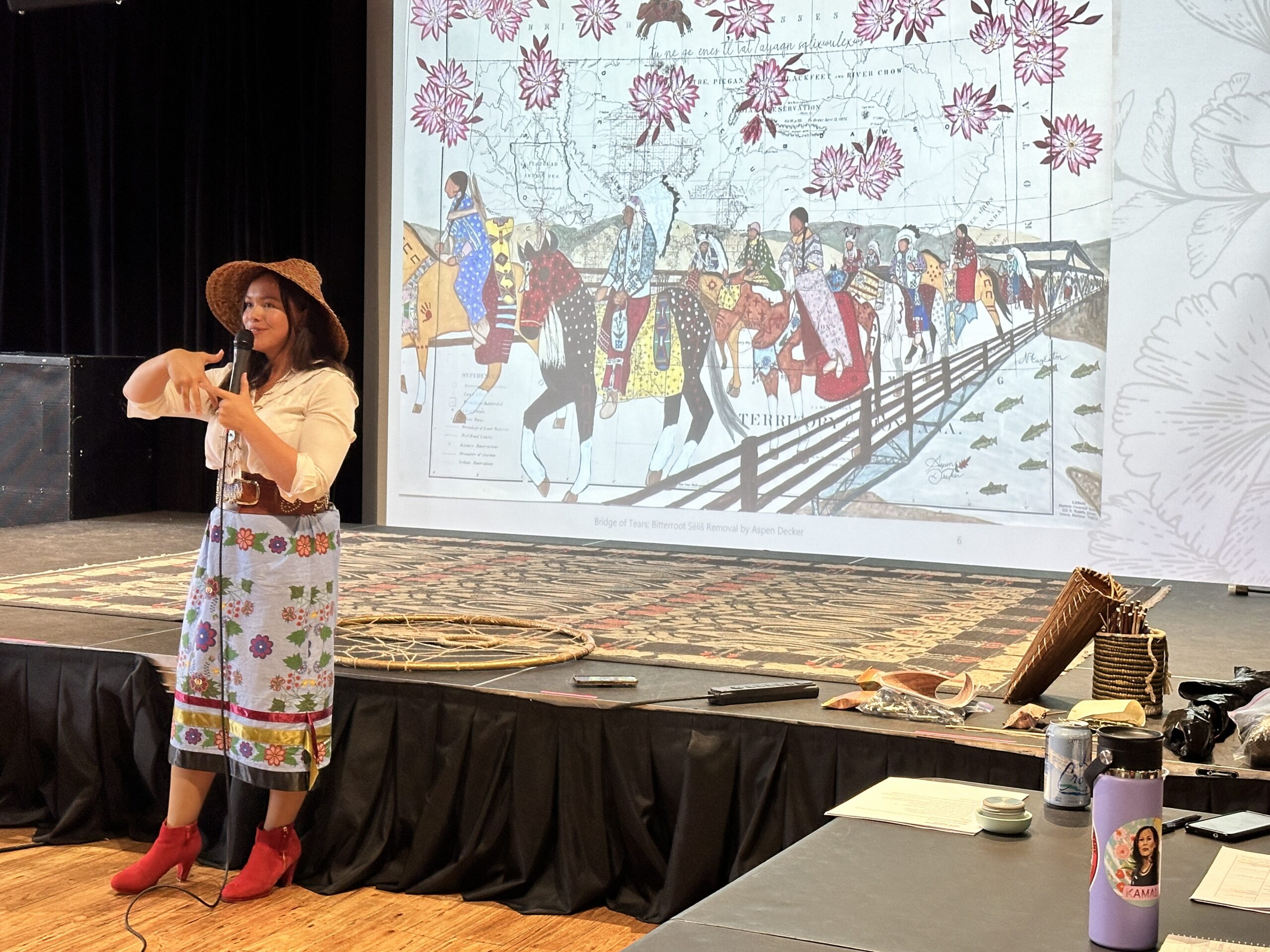 A person in a wide-brimmed hat, white blouse, floral skirt, and red boots presents on stage with a microphone. Behind them, a screen displays artwork titled 'Bridge of Tears: Bitterroot Salish Removal by Aspen Decker,' depicting people on horseback crossing a bridge with floral patterns and a map in the background. A woven circular object lies on the stage floor behind her.