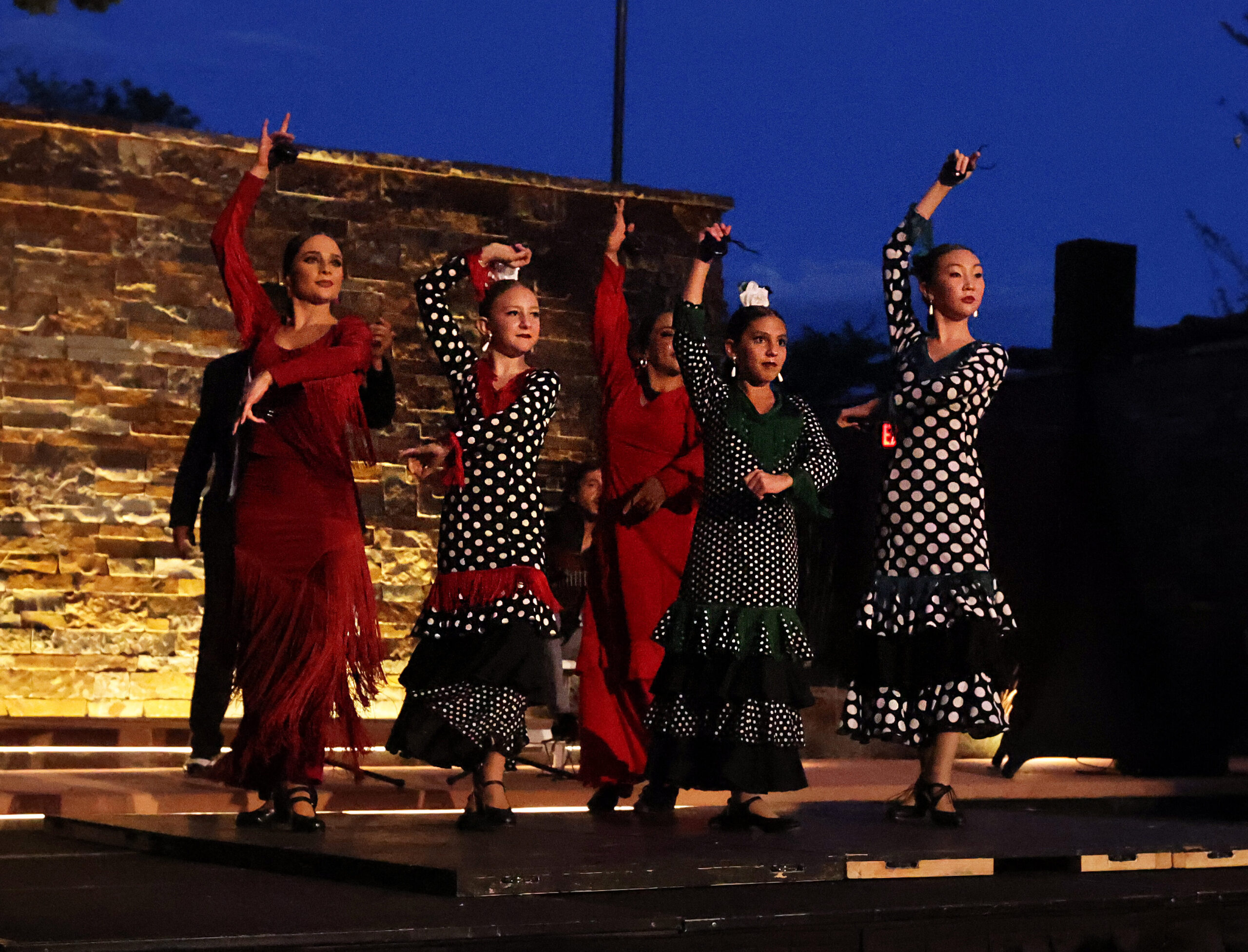 Five performers in traditional flamenco attire dance on an outdoor stage, wearing vibrant red, black, and white polka-dotted costumes with ruffles and fringes. The stage is set against a warmly lit stone wall, with string lights above and trees visible in the background, creating a festive atmosphere.