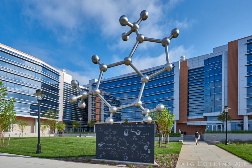 Large metallic molecular sculpture in front of a modern office building with reflective glass windows.