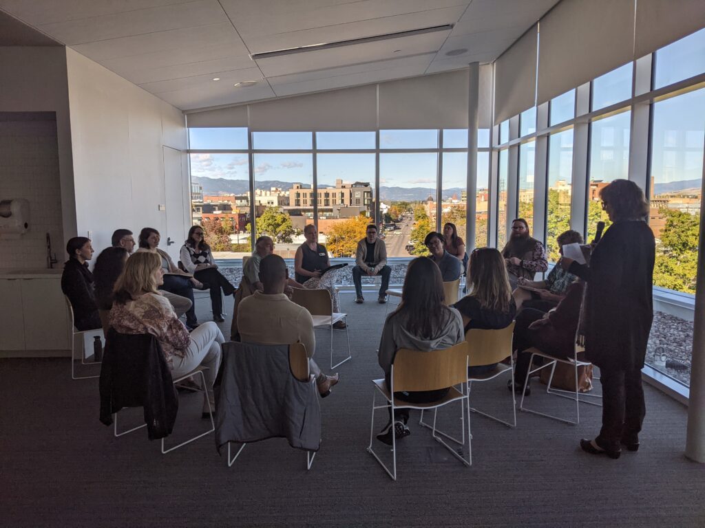 A group of people seated in a circle in a room with floor-to-ceiling windows where Missoula, Montana can be seen outside.